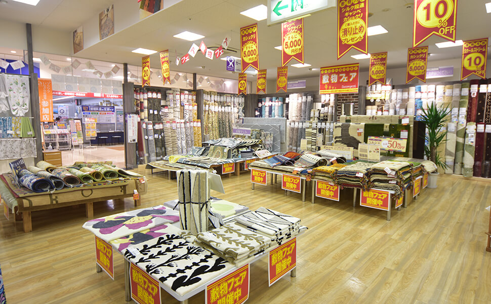 愛知県稲沢市に位置するカーテン専門店サンレジャン店内。絨毯フェアが開催中で赤と黄色の視認性の高いＰＯＰが並び、色々な絨毯が店頭に並ぶ。