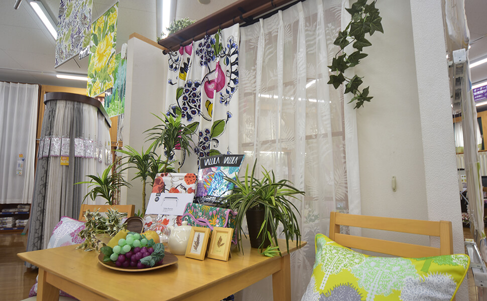 愛知県稲沢市に位置するカーテン専門店サンレジャン店内。テーブルの上には観葉植物。椅子の上には黄緑の幾何学模様が飾られたクッションが置かれる。カーテンにも、青や赤の花々がランダムに配置。