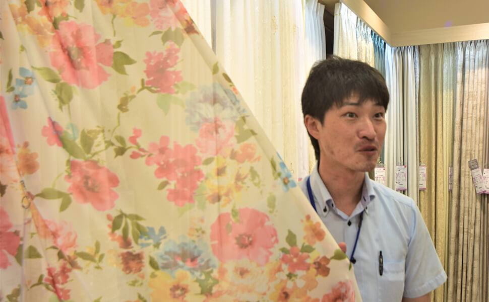 愛知県稲沢市に位置するカーテン専門店サンレジャン店内。カーテンを選びながら、おどけた表情を見せるサンレジャン店長。
