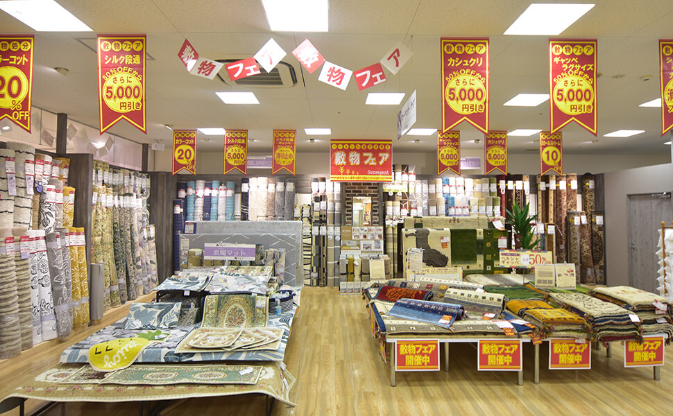 愛知県稲沢市に位置するカーテン専門店サンレジャン店内。稲沢店で開催中の絨毯フェアの売場。赤と黄色のＰＯＰが天井からぶら下げられ、様々なカラー模様のじゅうたんが所狭しと並ぶ。